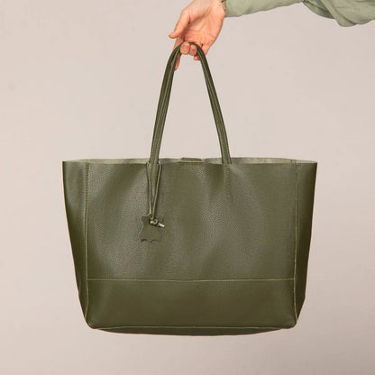 Eine Hand hält die grüne flyliving Umhängetasche "Susi" aus Leder, mit Doppelgriffen und einem kleinen Anhänger, vor einem schlichten beigen Hintergrund.