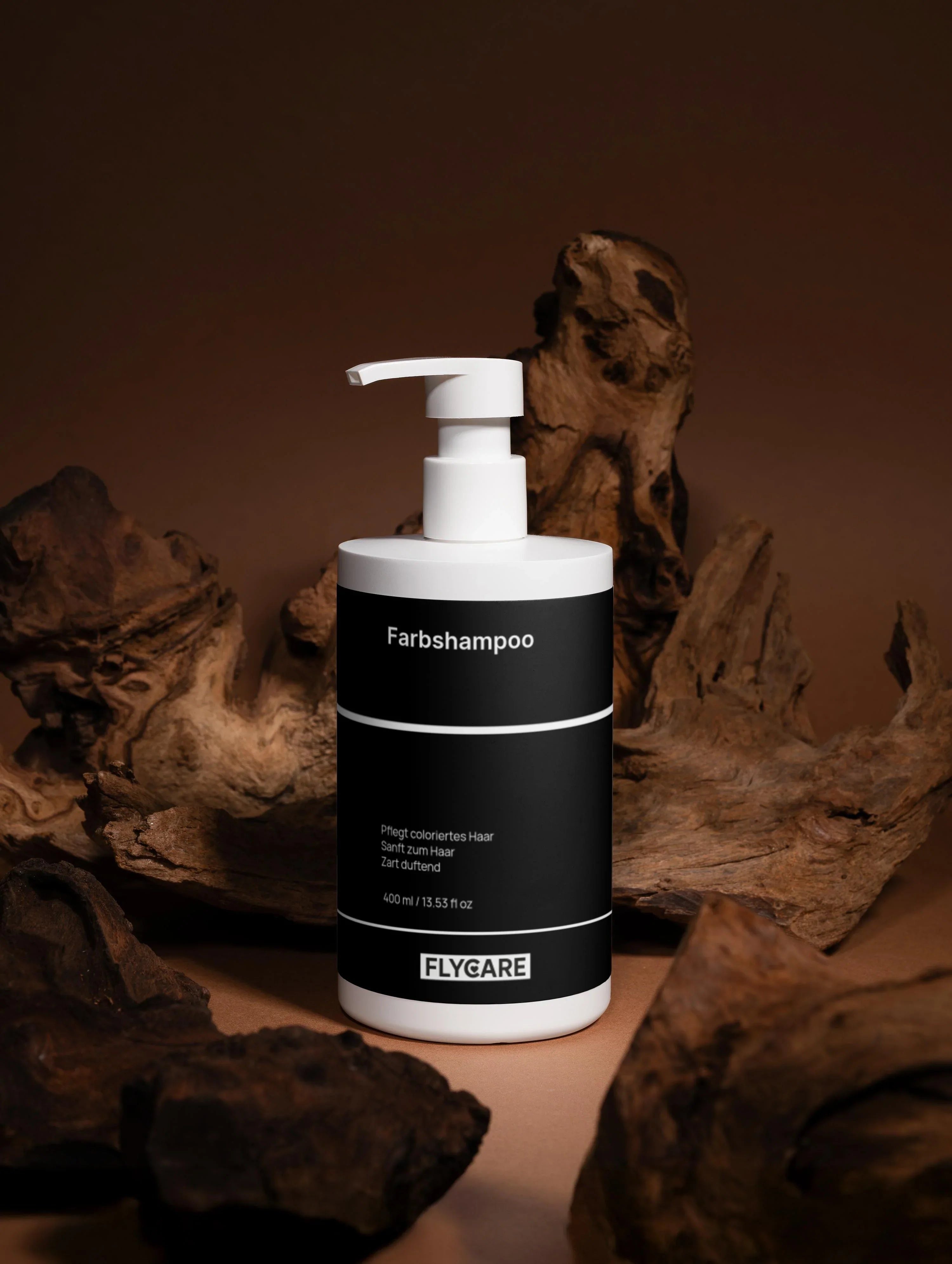 Farbshampoo von Flycare in einer weißen Flasche mit einer schwarzen Banderole, umgeben von Treibholz.
