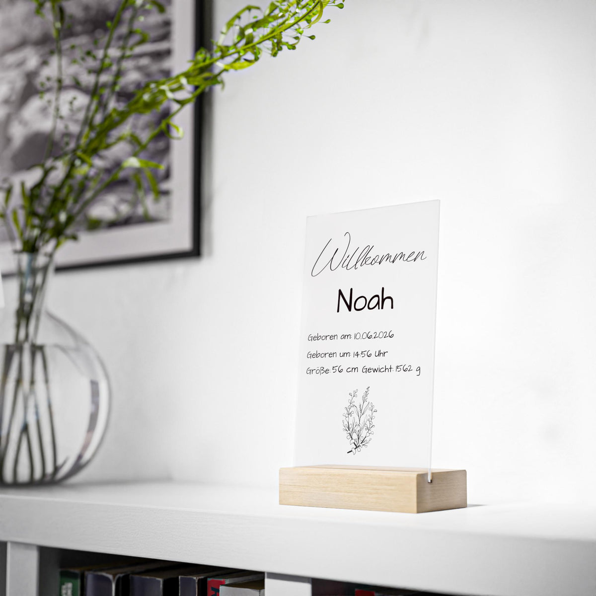 Weiße keramikvase mit grünem monstera-blatt und beigem pampasgras neben personalisierter klarer acryl-tafel geburt mit holzsockel