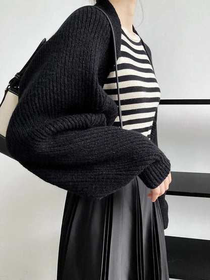 Eine Person trägt den Touka Strick Shrug in Schwarz von marigoldshadows über einem schwarz-weiß gestreiften Top und plissierten Rock, hält eine schwarz-weiße Tasche und steht in minimalistischer Umgebung.