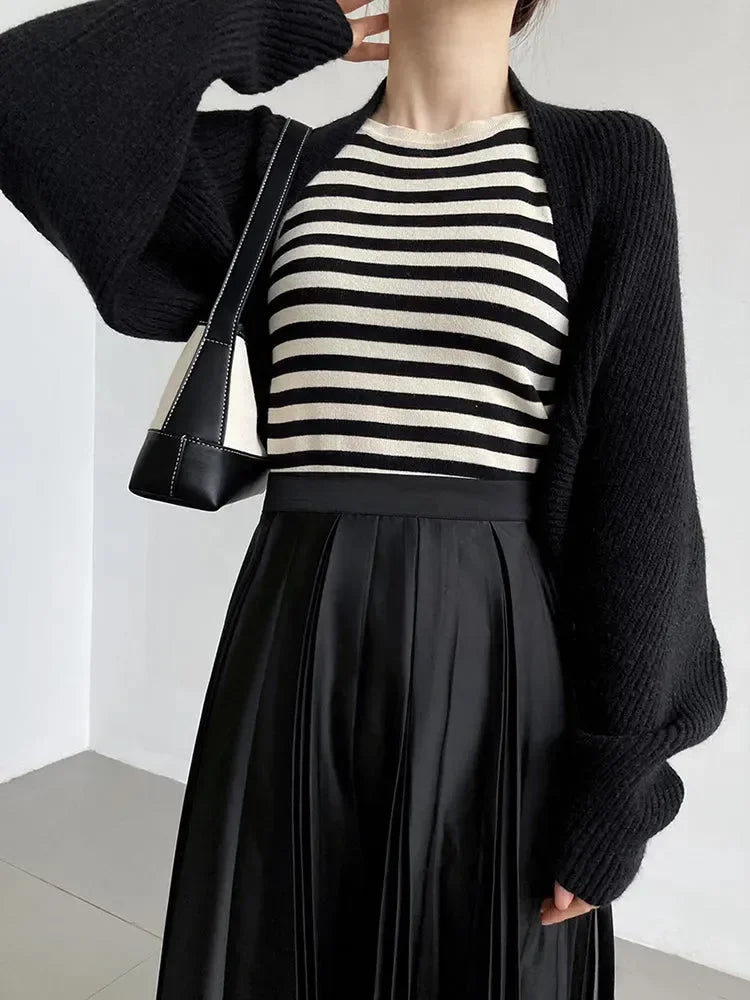 Eine Person trägt Damen Mode: schwarze Oberteile, darunter den Touka Strick Shrug in Schwarz von marigoldshadows, eine schwarz-weiß gestreifte Bluse, einen plissierten Rock und eine schwarze Umhängetasche. Der Kopf ist nicht sichtbar.