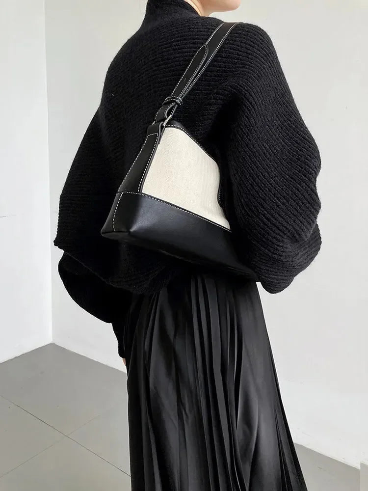 Eine Person, die den marigoldshadows Touka Strick Shrug in Schwarz trägt, gestylt mit einem Faltenrock und einer zweifarbigen Umhängetasche, steht vor einer schlichten, hellen Wand - ein schicker Damen Mode-Layering Inspo. Gesicht nicht sichtbar.