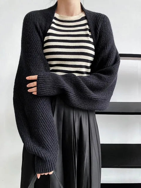 Eine Frau modelliert die marigoldshadows Touka Strick Shrug in schwarz über einem schwarz-weiß gestreiften Oberteil und einem schwarzen Faltenrock und hebt damit elegante Damen Mode vor einem schlichten weißen Hintergrund hervor.
