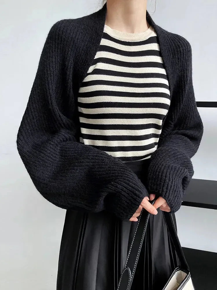 Eine Frau modelliert Damen Mode, trägt ein schwarz-weiß gestreiftes Oberteil, die marigoldshadows Touka Strick Shrug in schwarz und einen schwarzen Rock, während sie eine passende Handtasche hält. Nur ihr Oberkörper ist vor einem einfarbigen Hintergrund zu sehen.