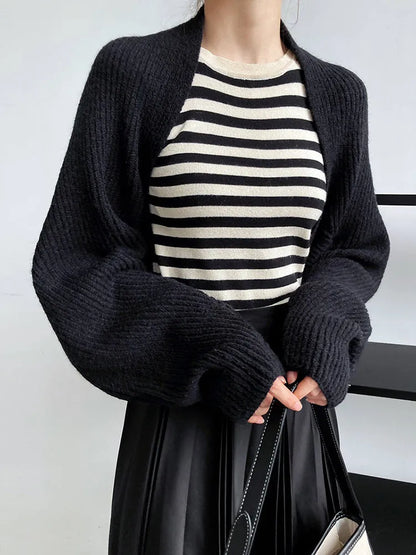 Eine Frau modelliert Damen Mode, trägt ein schwarz-weiß gestreiftes Oberteil, die marigoldshadows Touka Strick Shrug in schwarz und einen schwarzen Rock, während sie eine passende Handtasche hält. Nur ihr Oberkörper ist vor einem einfarbigen Hintergrund zu sehen.