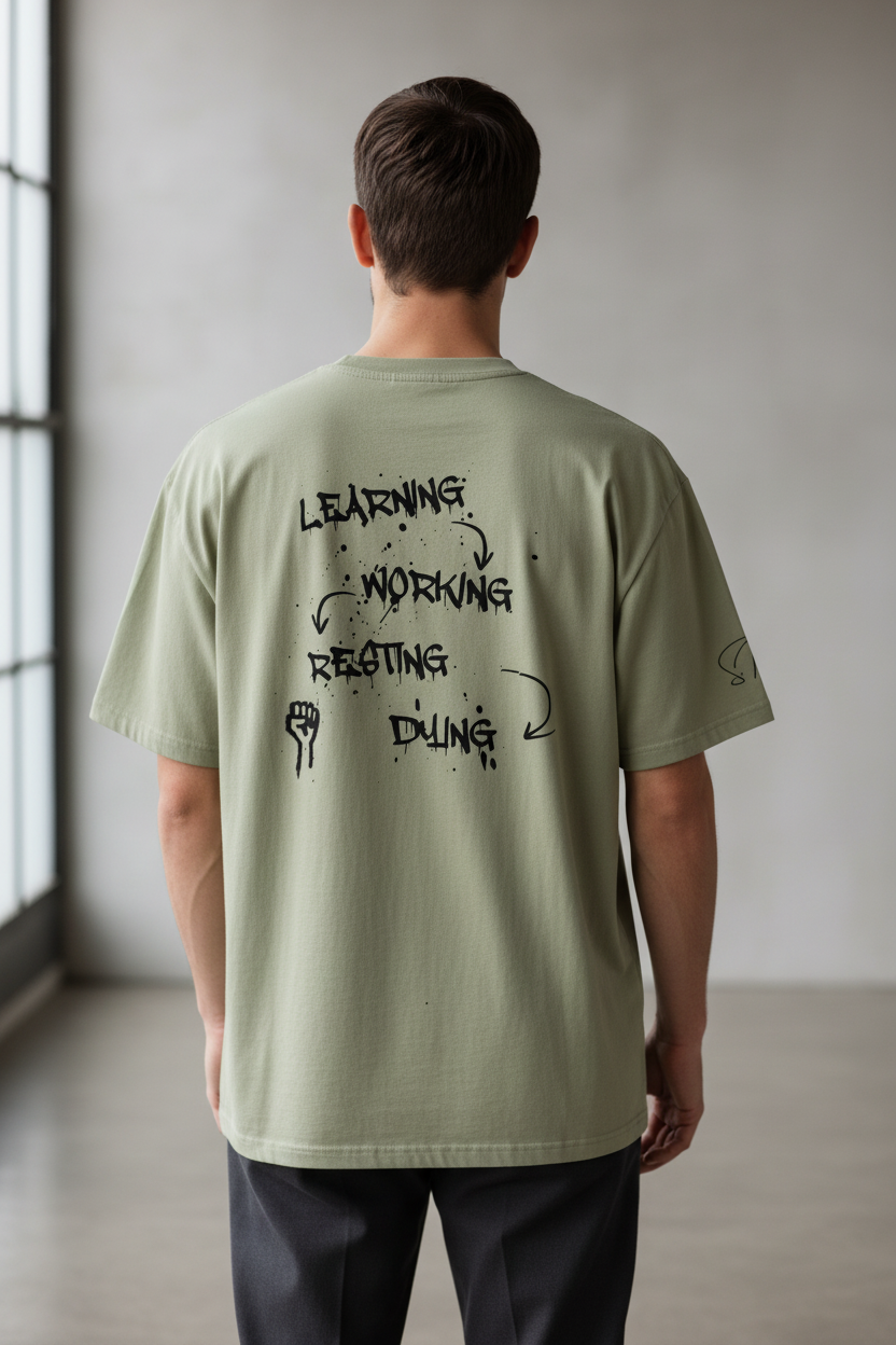 Oversized sage green heavy cotton T-shirt by flyliving, back view, with a black graffiti graphic of 'LEARNING-WORKING-RESTING-DYING' cycle and a fist.