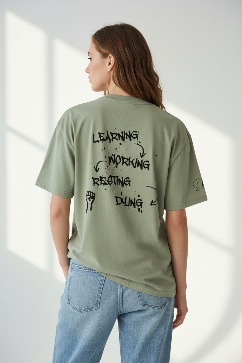 Muted green heavy cotton oversized T-shirt by flyliving, with a washed 90s-look and a back print featuring the LEARNING-WORKING-RESTING-DYING cycle, worn with blue jeans.