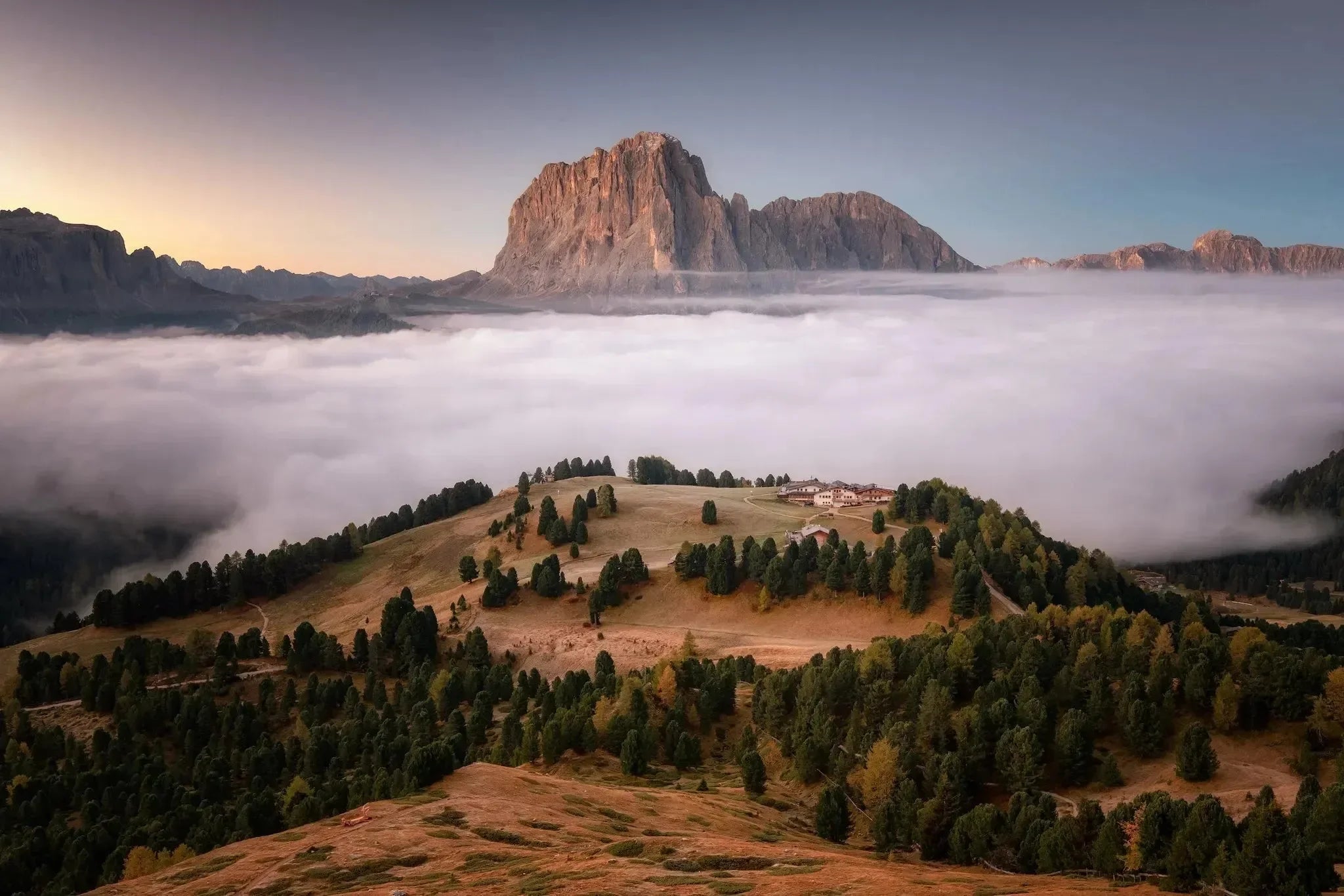 Sanfte Hügel und Baumgruppen umgeben ein Dorf, während dichter Nebel vorbeizieht, mit einem dramatischen Berg unter klarem Himmel - eine perfekte Szene für Wanderlust Alpen von gestalten EU Shop, ideal für Alpen Abenteuer oder Wanderwege Alpen.