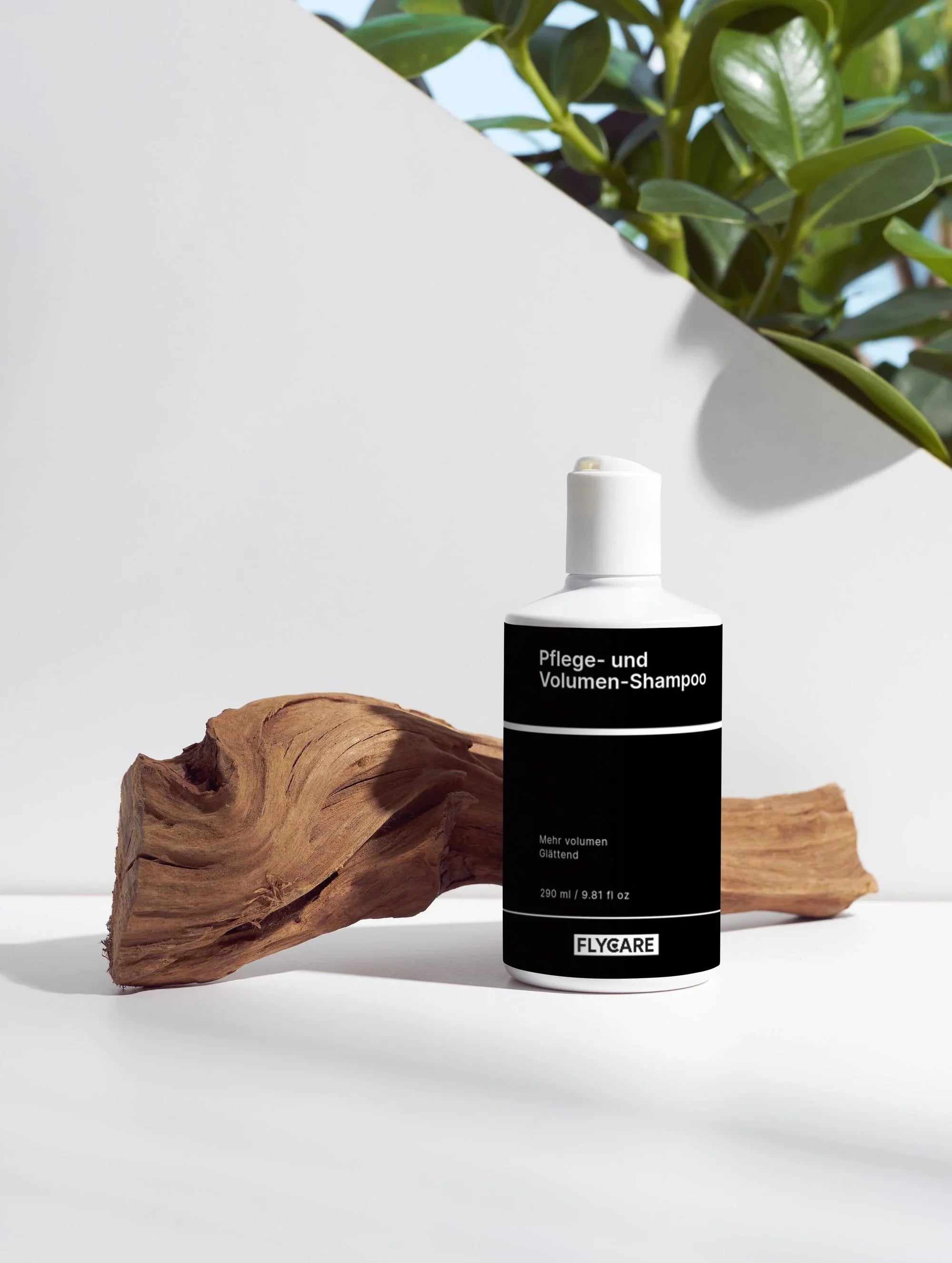 Pflege- und Volumen-Shampoo von Flycare, neben einem Stück Treibholz auf weißem Untergrund, mit grünen Pflanzen im Hintergrund.
