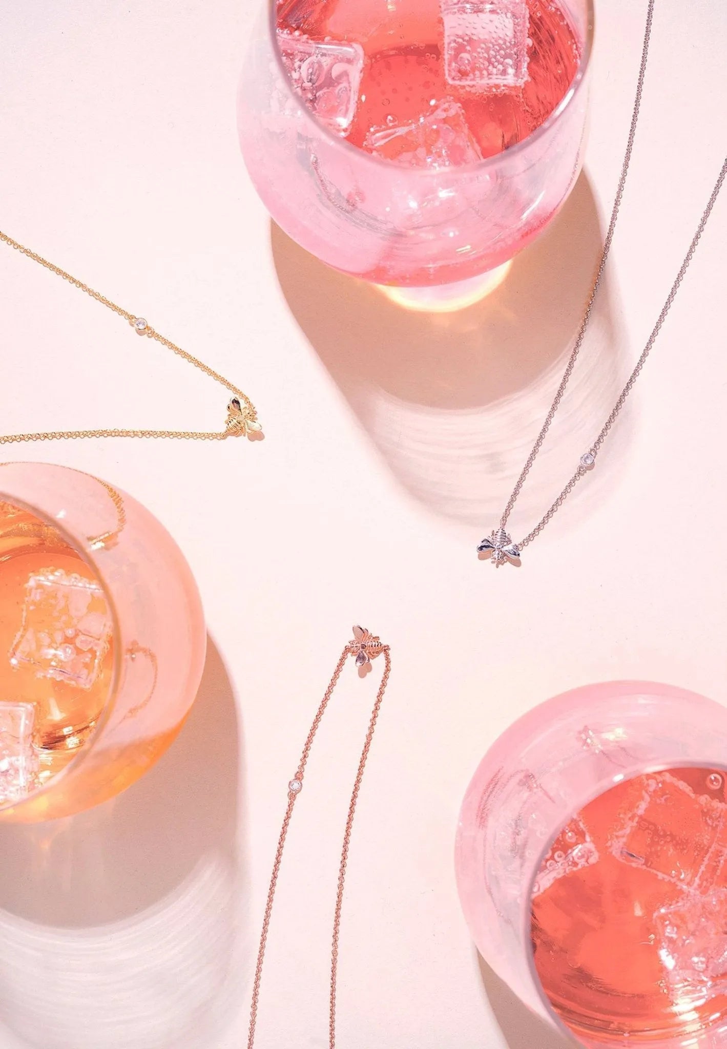 Drei rosafarbene Cocktails mit Eiswürfeln ruhen auf einer hellen Fläche, akzentuiert durch zarte Goldketten von LATELITA, darunter die elegante, vergoldete Halskette "Kunigunde". Helles Licht wirft weiche Schatten und reizvolle Reflexe auf die Szene.