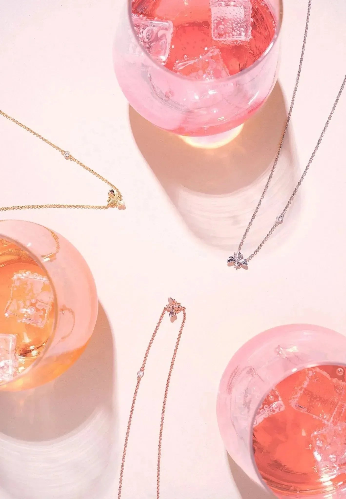 Drei rosafarbene Cocktails mit Eiswürfeln ruhen auf einer hellen Fläche, akzentuiert durch zarte Goldketten von LATELITA, darunter die elegante, vergoldete Halskette "Kunigunde". Helles Licht wirft weiche Schatten und reizvolle Reflexe auf die Szene.