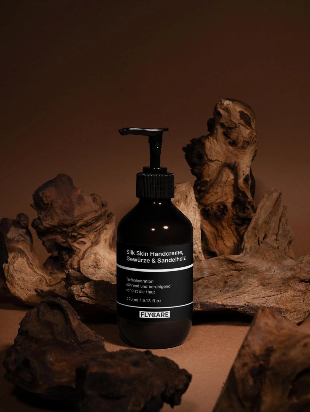 Seifenspender mit Handcreme, Seide, Gewürze und Sandelholz von Flycare, vor Treibholz in dunklen Farbtönen.
