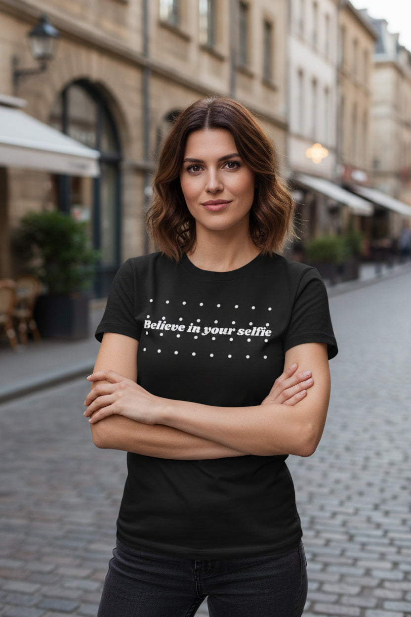 Black cotton T-shirt by flyliving with "Believe in your selfie" text and white dots, worn by a woman with crossed arms outdoors.