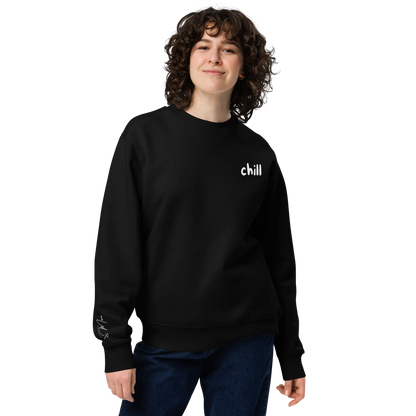 Eine Person mit lockigem Haar lächelt, sie trägt den schwarzen flyliving Pullover "chill" aus 100% organischer Baumwolle und eine blaue Jeans, vor einem schlichten Hintergrund - ideal für einen entspannten Lifestyle.