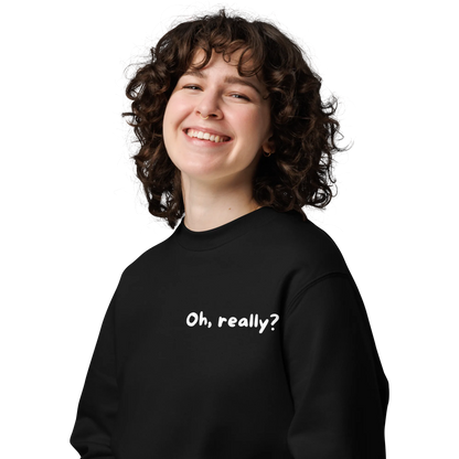 Eine Person mit lockigem Haar lächelt breit und trägt den flyliving Pullover "Oh, really?" mit weißem Text auf der Vorderseite. Der Hintergrund ist einfarbig und dunkel.