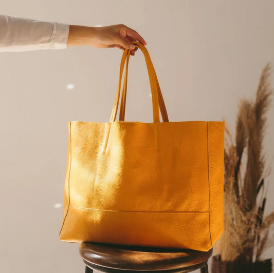 Jemand hält die flyliving Umhängetasche "Susi" aus gelbem Leder an ihren Griffen über einem dunklen Holzhocker, während getrocknete Pflanzen und sanftes Sonnenlicht das luxuriöse Material im Hintergrund hervorheben.